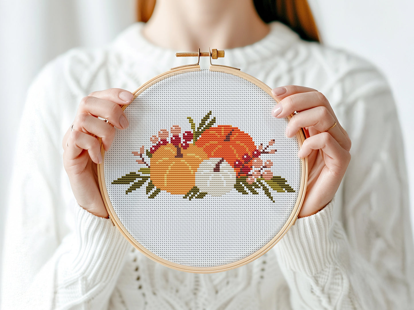 Person holding a cross-stitched hoop with autumn-themed design featuring pumpkins and leaves.
