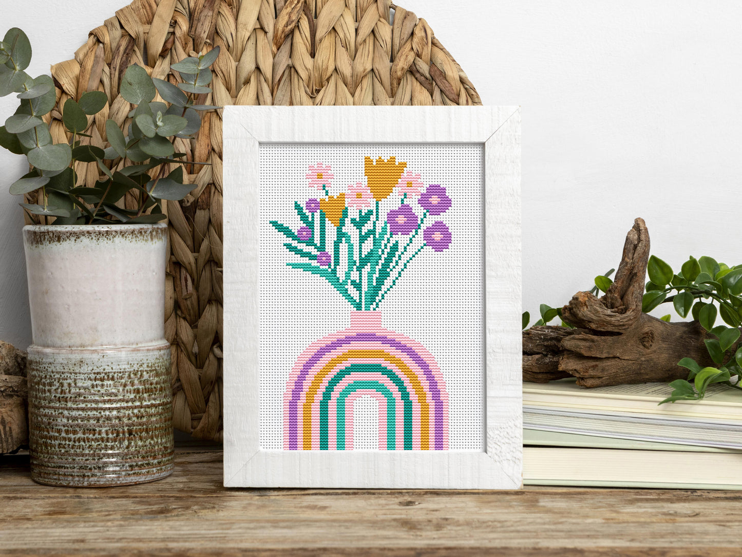 Framed cross-stitched artwork with flowers and a rainbow on a wooden surface.