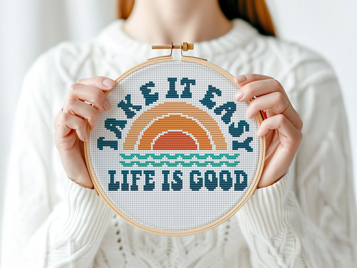 Person holding a cross-stitched hoop with 'Take It Easy, Life Is Good' text and sunset design.