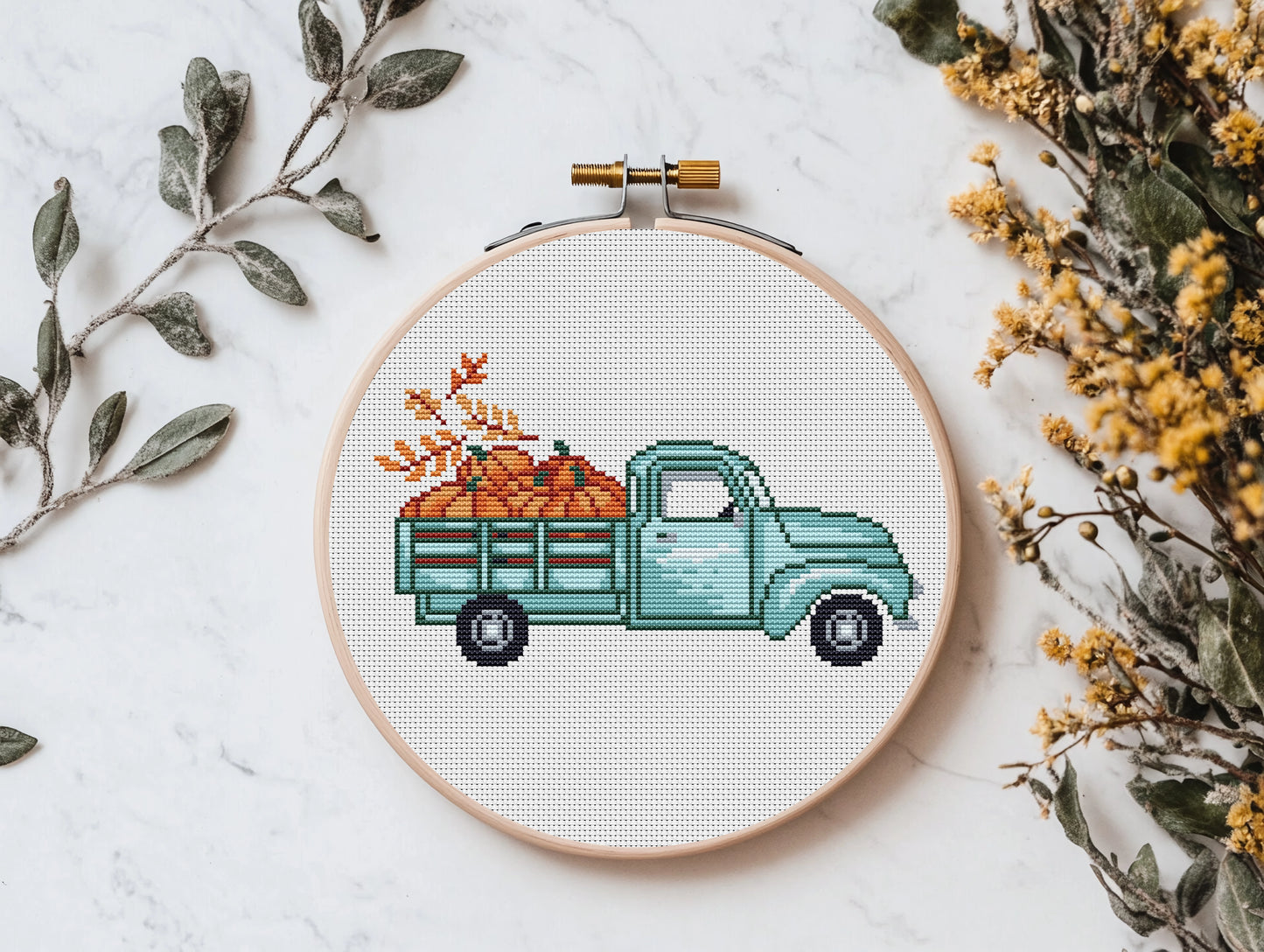 Cross-stitched design of a blue truck with pumpkins in an embroidery hoop, surrounded by dried leaves and flowers on a light background.