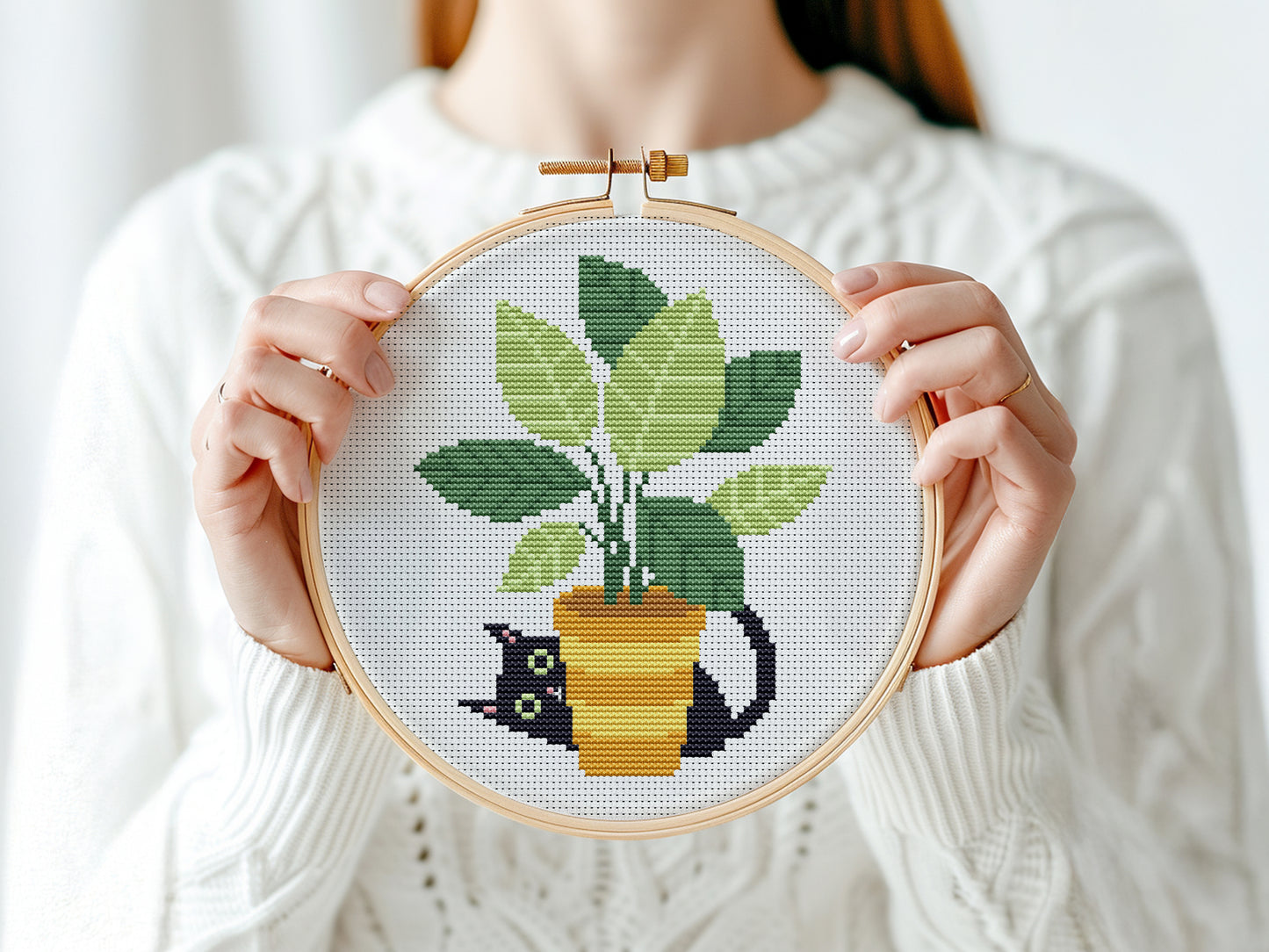 Person holding a cross-stitched plant and cat design in an embroidery hoop.