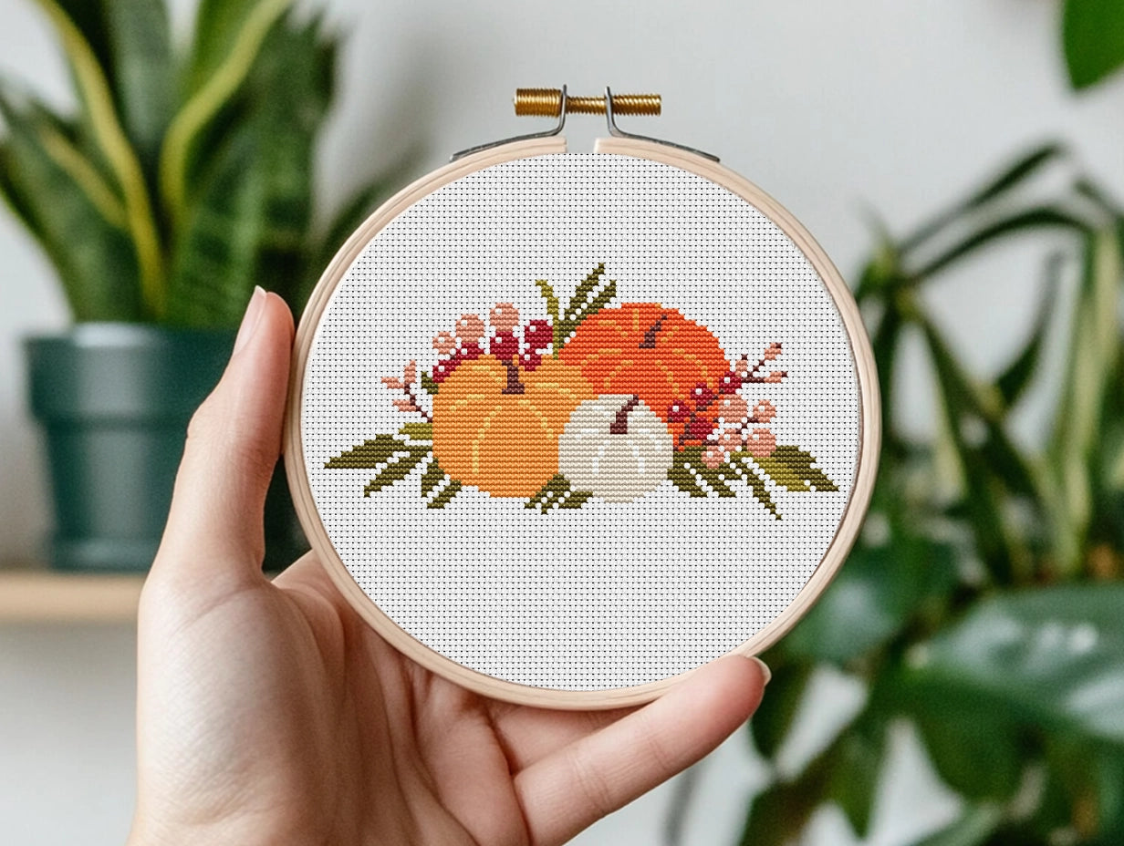 Hand holding a cross-stitched hoop with a pumpkin design against a plant background