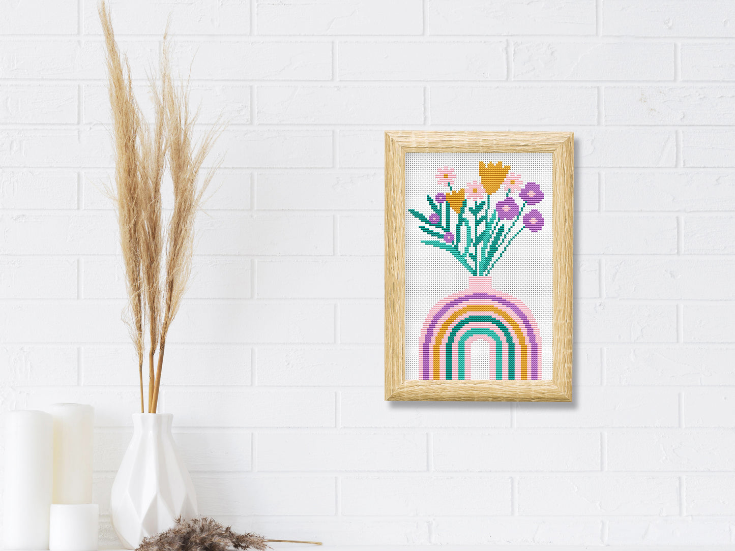 Framed cross-stitched artwork with flowers and a rainbow on a white tiled wall.