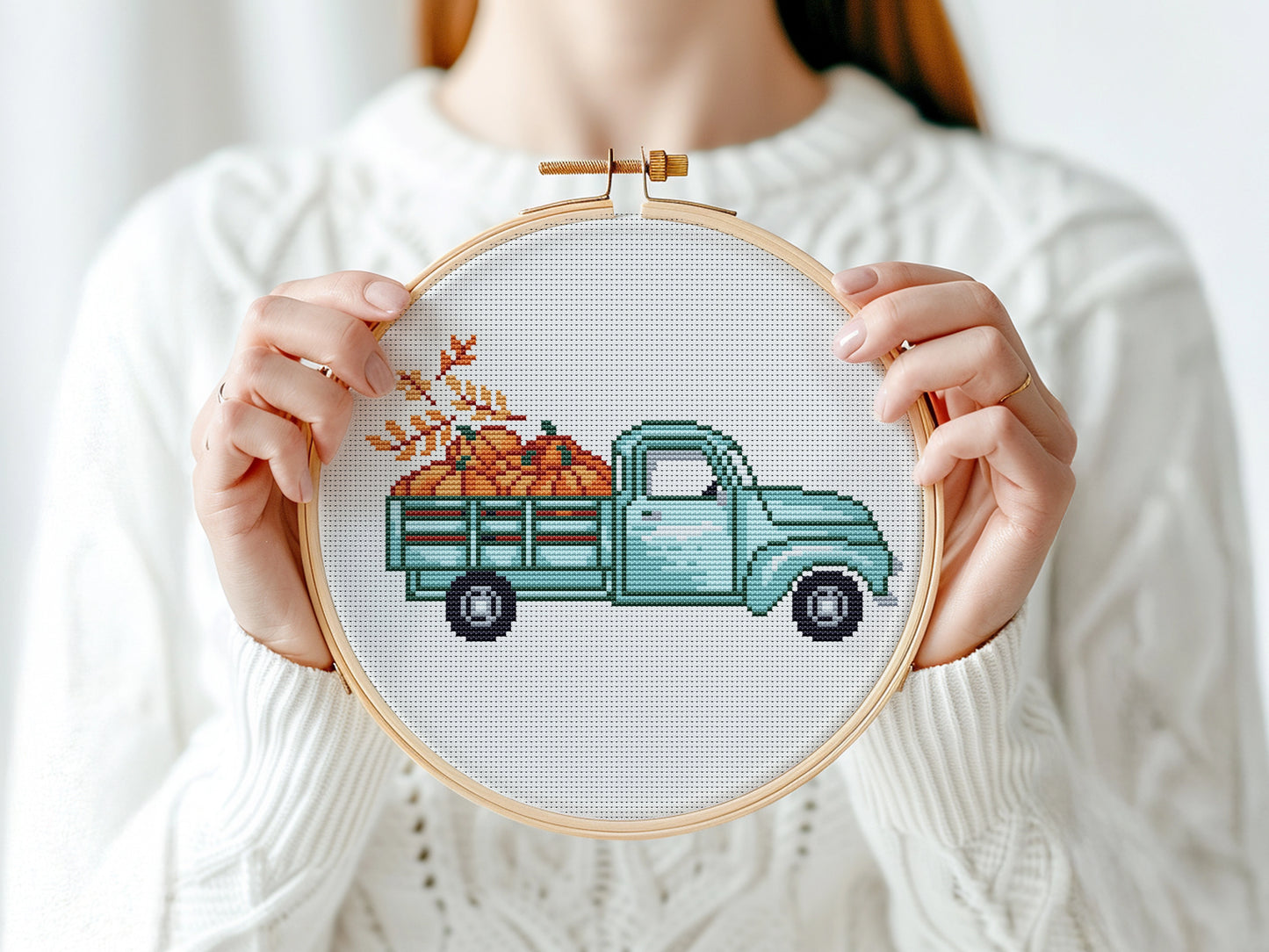 Person holding a cross-stitched hoop with a teal truck and pumpkins design.
