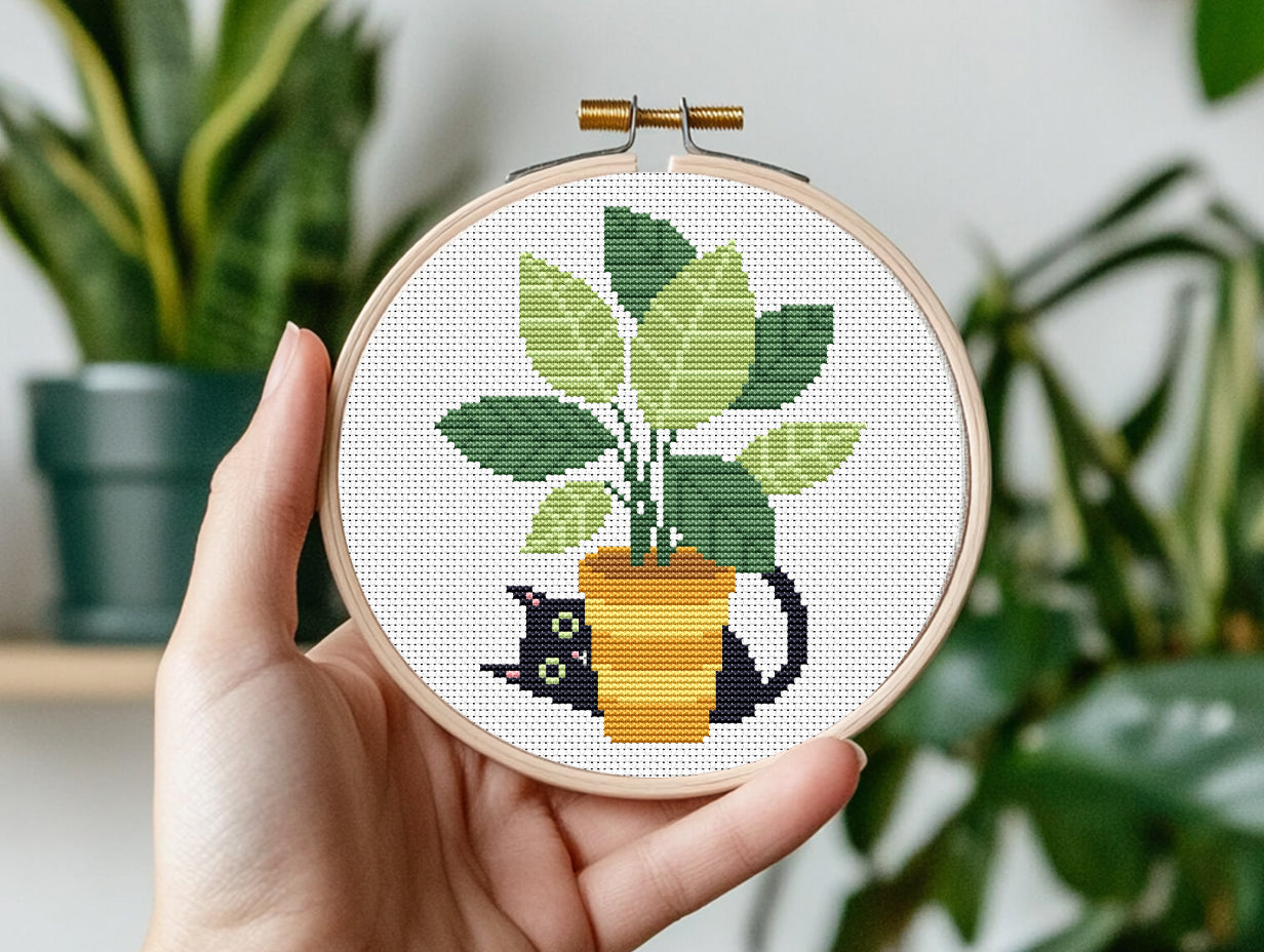 Hand holding a cross-stitched hoop with a potted plant design, surrounded by green plants.