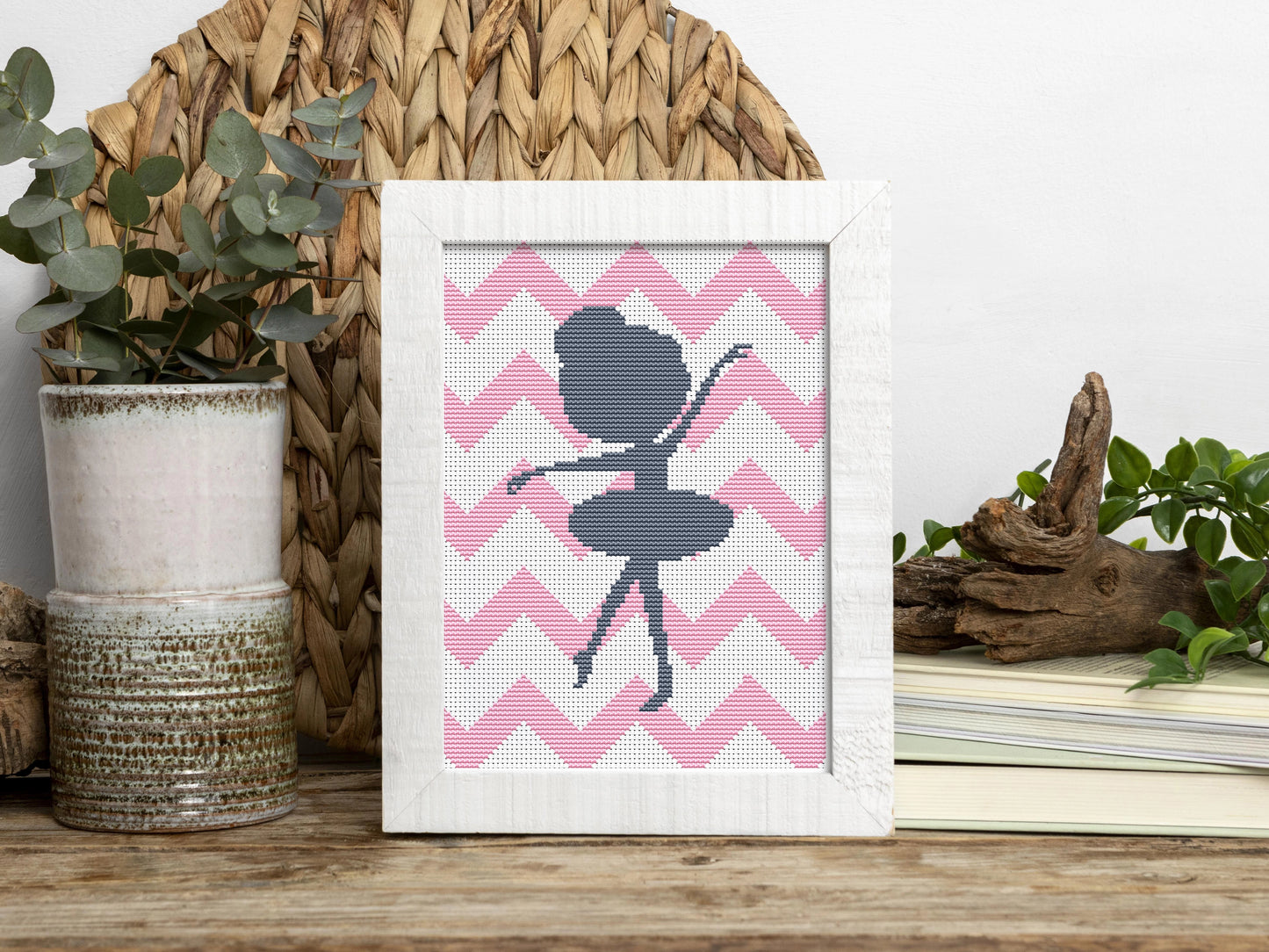Framed cross-stitched art of a ballerina on pink chevron pattern with decorative items in the background.