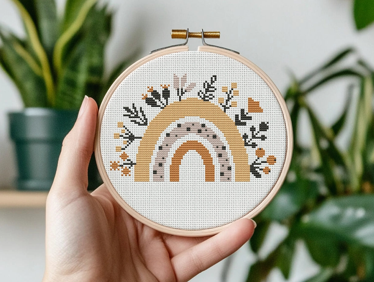 Hand holding a cross-stitched hoop with a rainbow design against a blurred plant background