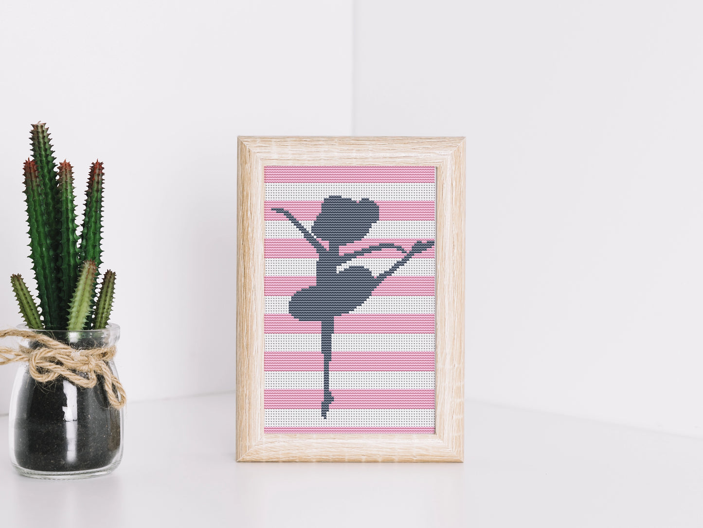 Framed cross-stitched art of a ballerina on a pink and white striped background, next to a potted plant.