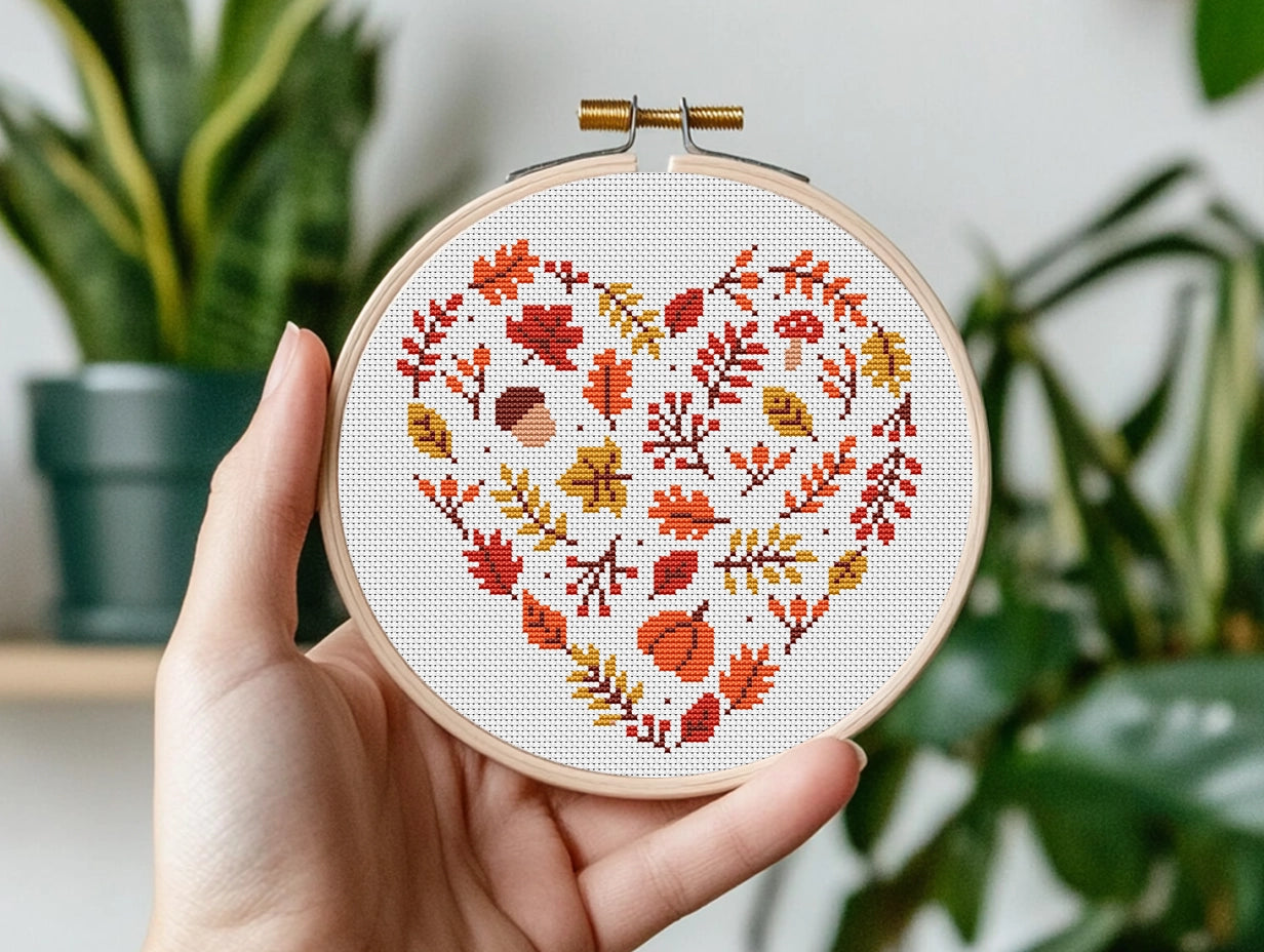 Hand holding a cross-stitched hoop with autumn leaf pattern against a blurred indoor plant background