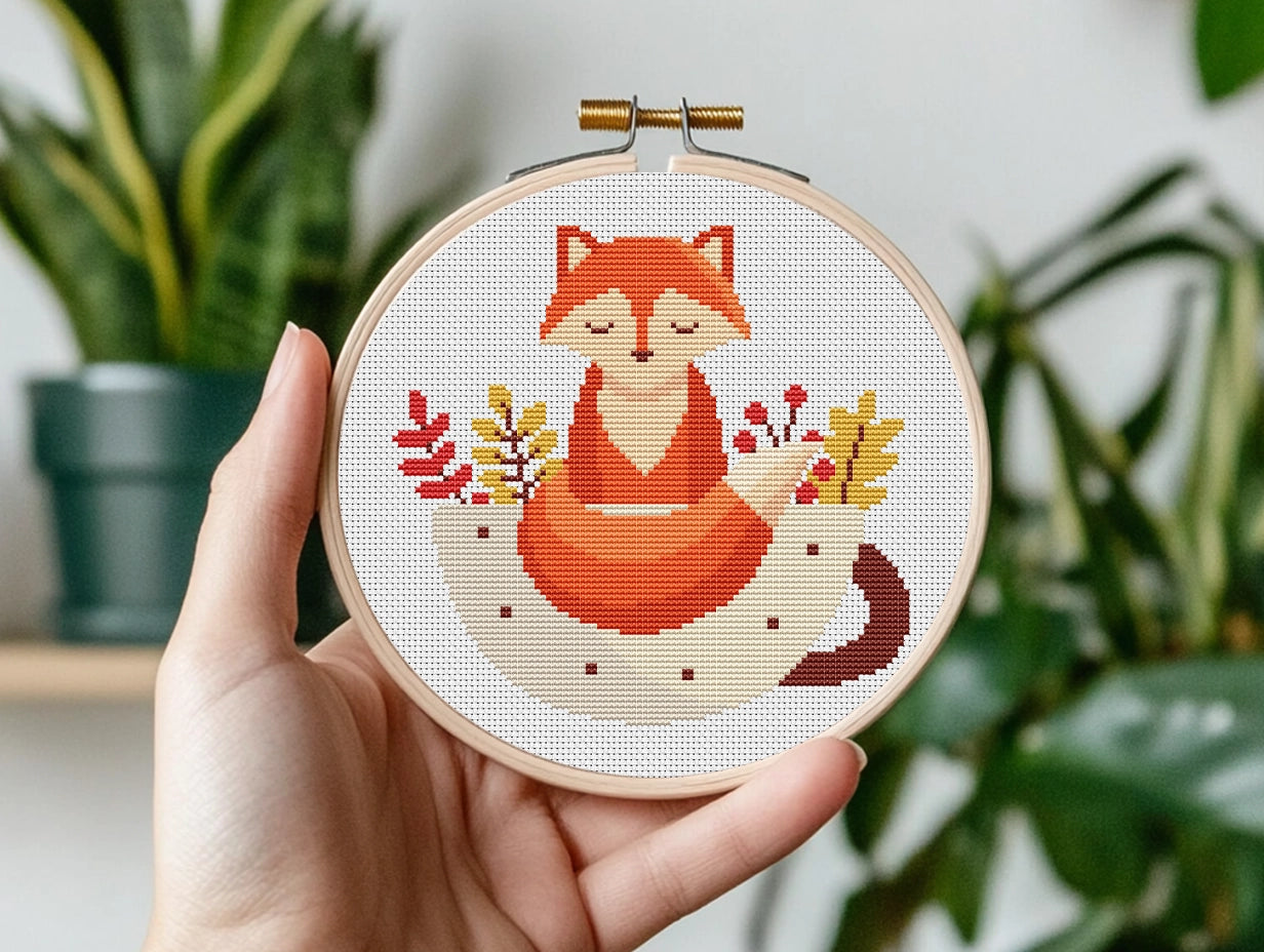 Hand holding a cross-stitched hoop with a fox design against a plant background