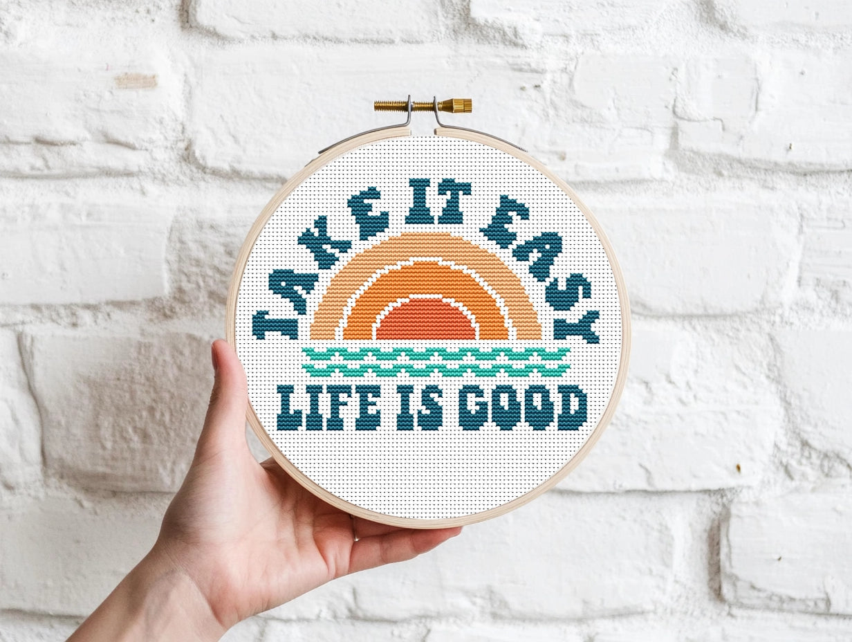 Embroidery hoop with 'Take it easy, life is good' text against a white brick wall.