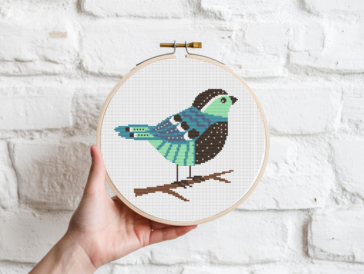 Embroidery hoop with a colorful bird design held against a white brick wall.