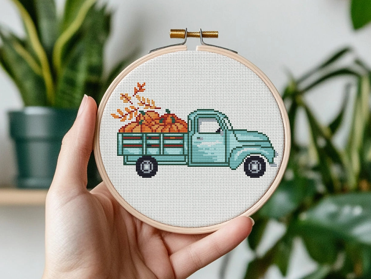 Hand holding a cross-stitched hoop with a blue truck and pumpkins design, against a blurred indoor plant background.