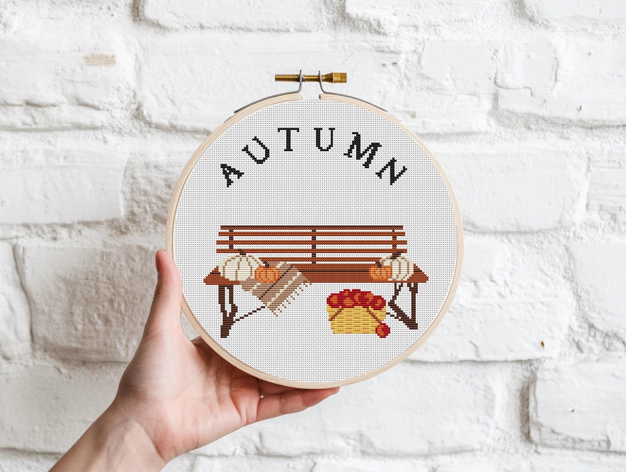 Cross-stitched hoop with 'AUTUMN' and bench design held against a white brick wall.