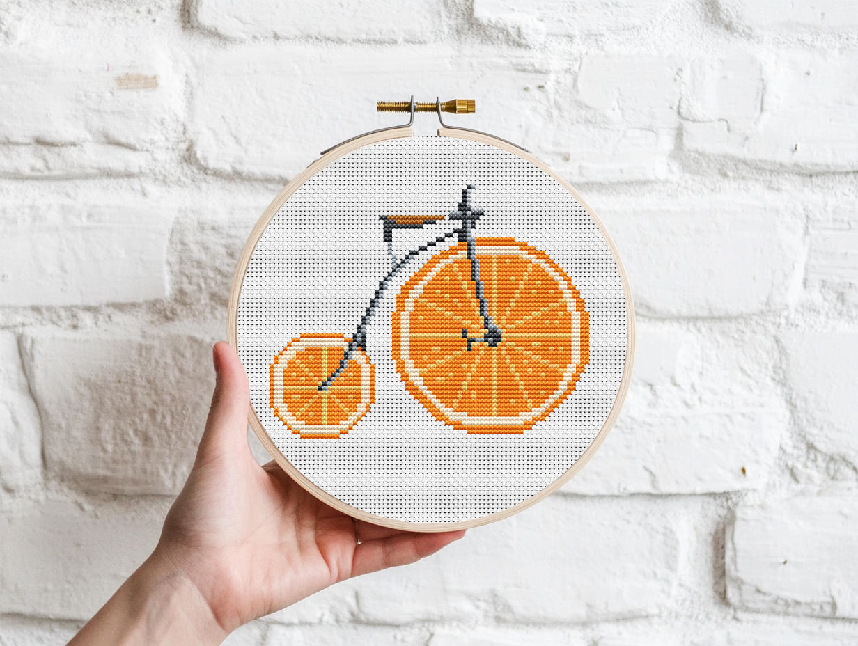 Embroidery hoop with a bicycle design made of orange shapes held against a white brick wall.