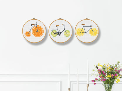 Three embroidered hoop art pieces with bicycles on oranges, lemons, and limes against a white wall.