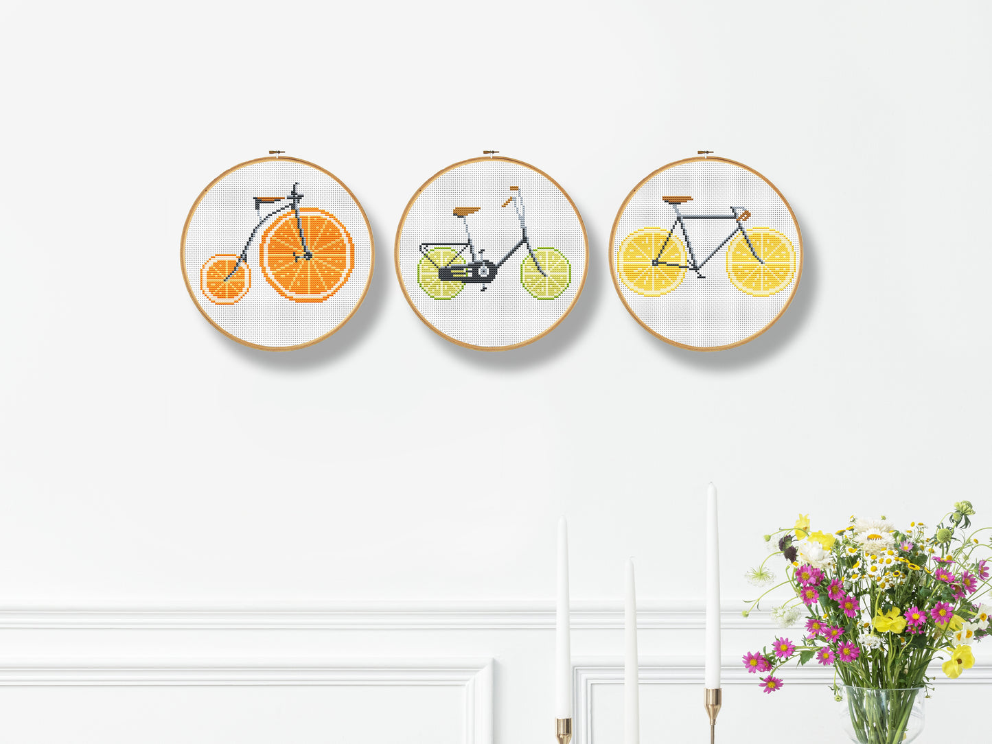 Three embroidered hoop art pieces with bicycles on oranges, lemons, and limes against a white wall.
