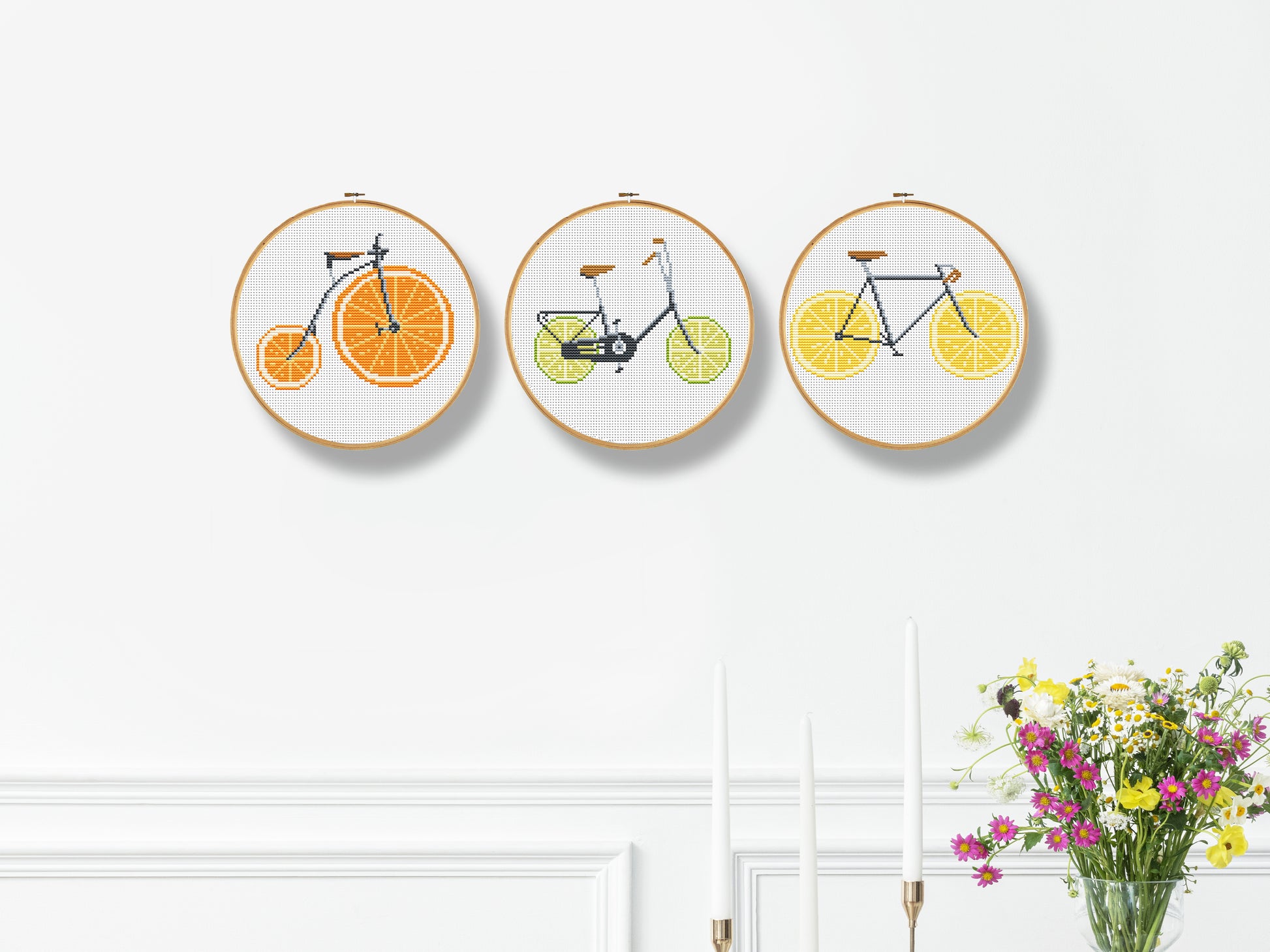 Three embroidered hoop art pieces with bicycles on oranges, lemons, and limes against a white wall.