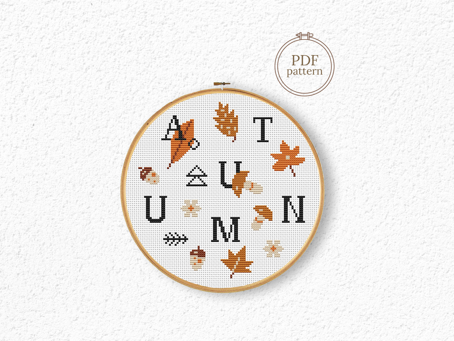 Cross-stitched hoop with 'AUTUMN' text and leaf patterns on a textured white background