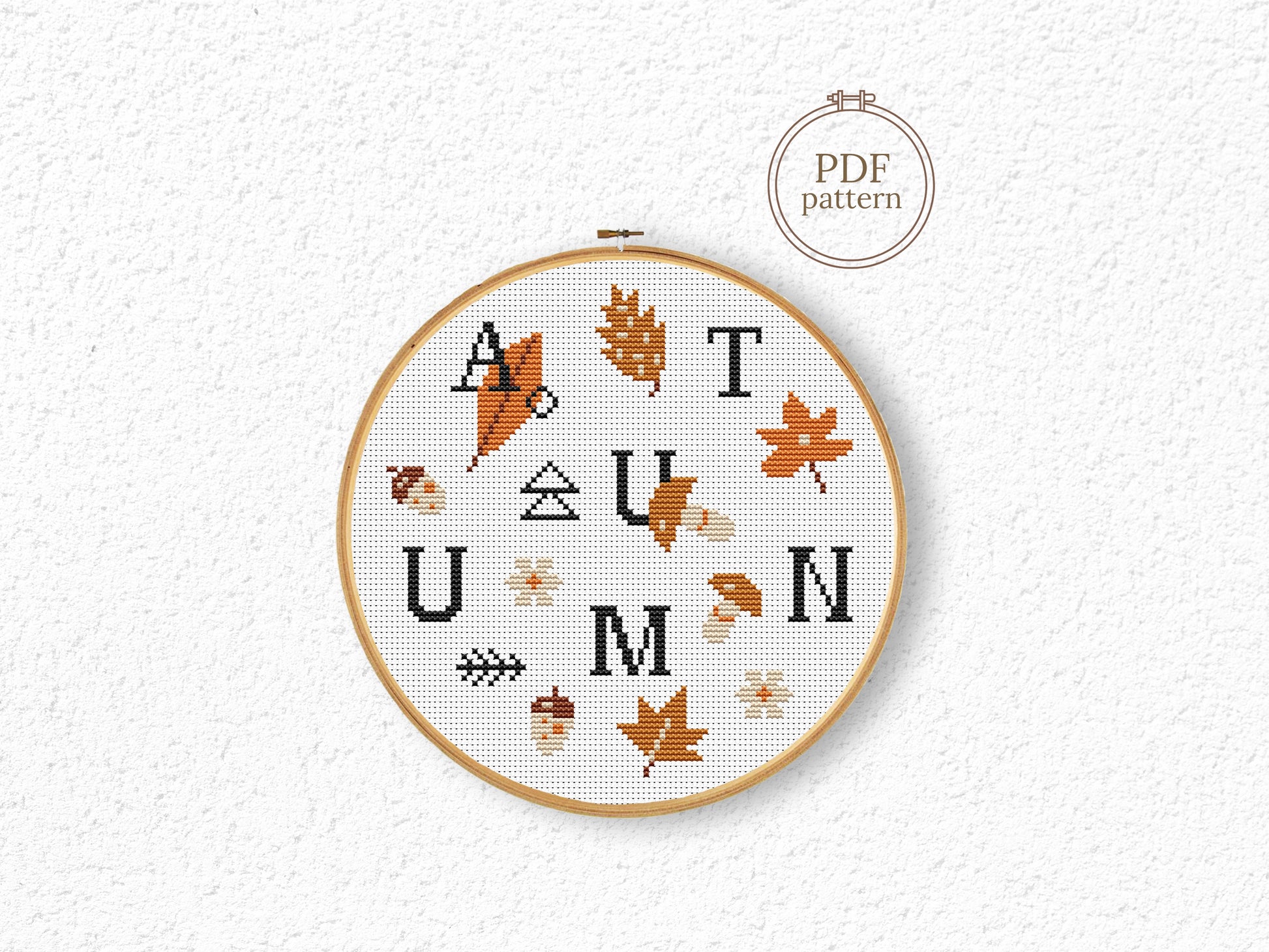 Cross-stitched hoop with 'AUTUMN' text and leaf patterns on a textured white background