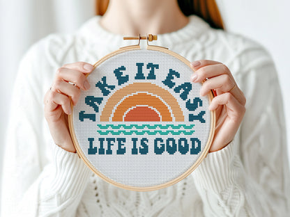 Person holding a cross-stitched hoop with 'Take It Easy, Life Is Good' text and sunset design.