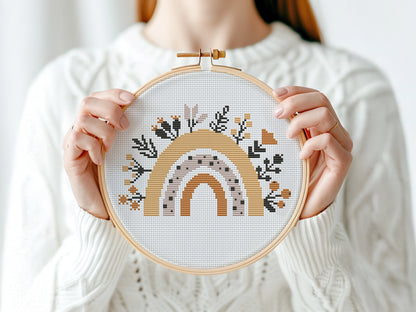 Person holding a cross-stitched hoop with a rainbow design against a white background
