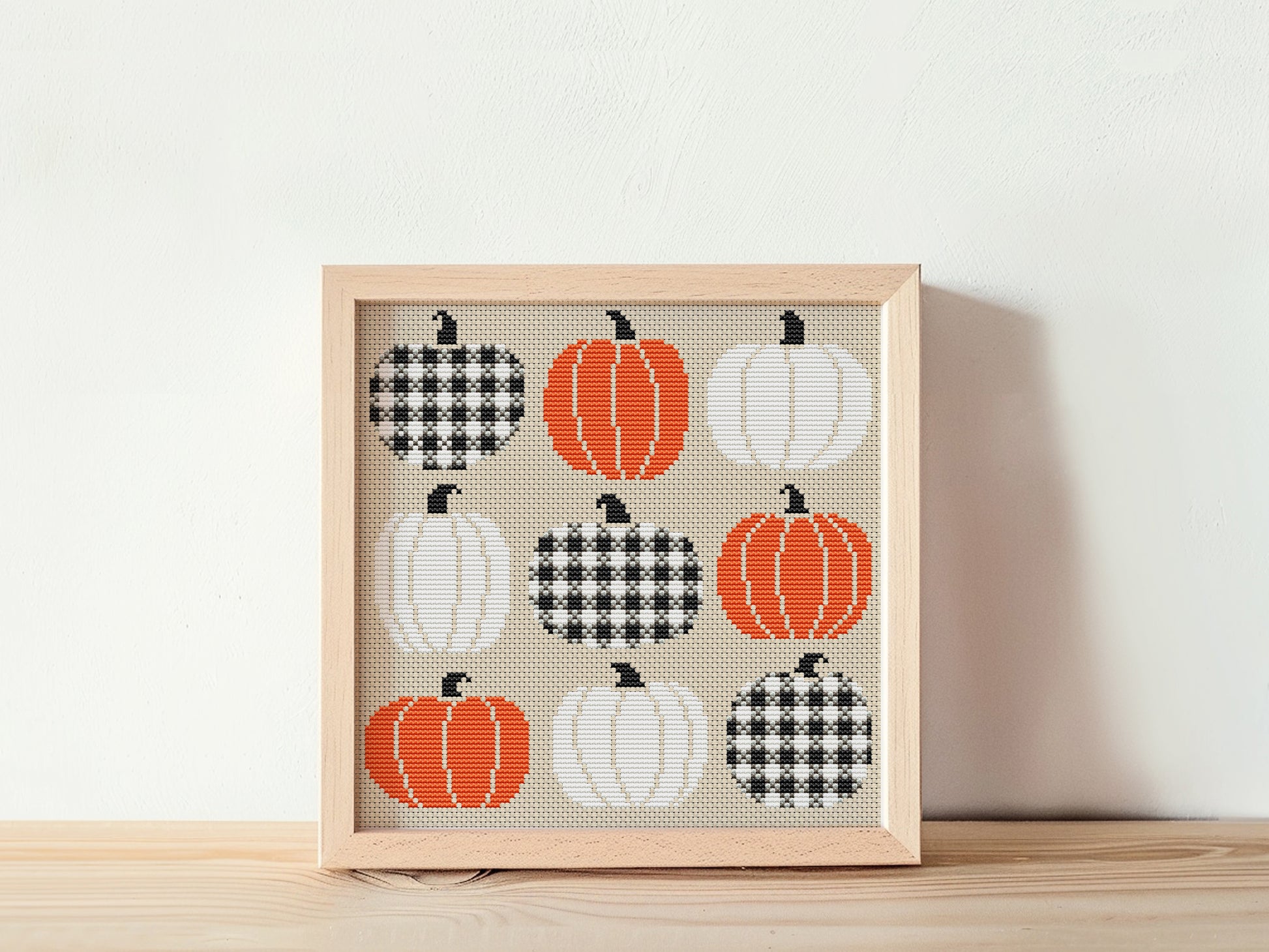 Framed cross-stitch art with pumpkins on a beige background