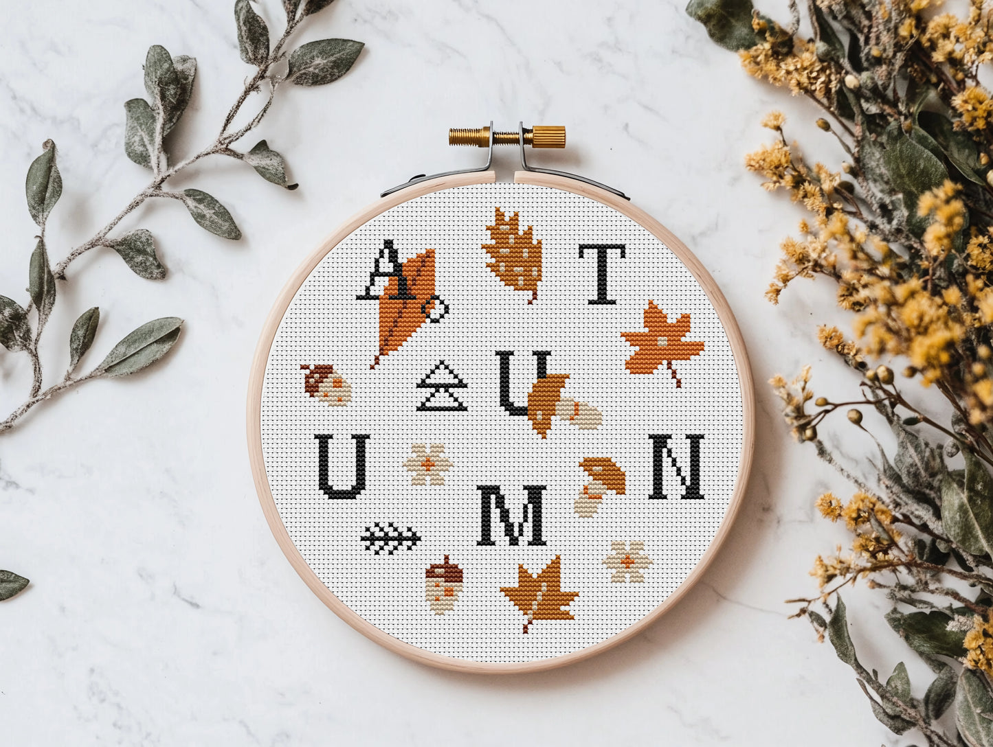 Cross-stitched autumn design in an embroidery hoop with leaves and acorns on a white background.