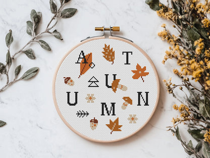 Cross-stitched autumn design in an embroidery hoop with leaves and acorns on a white background.