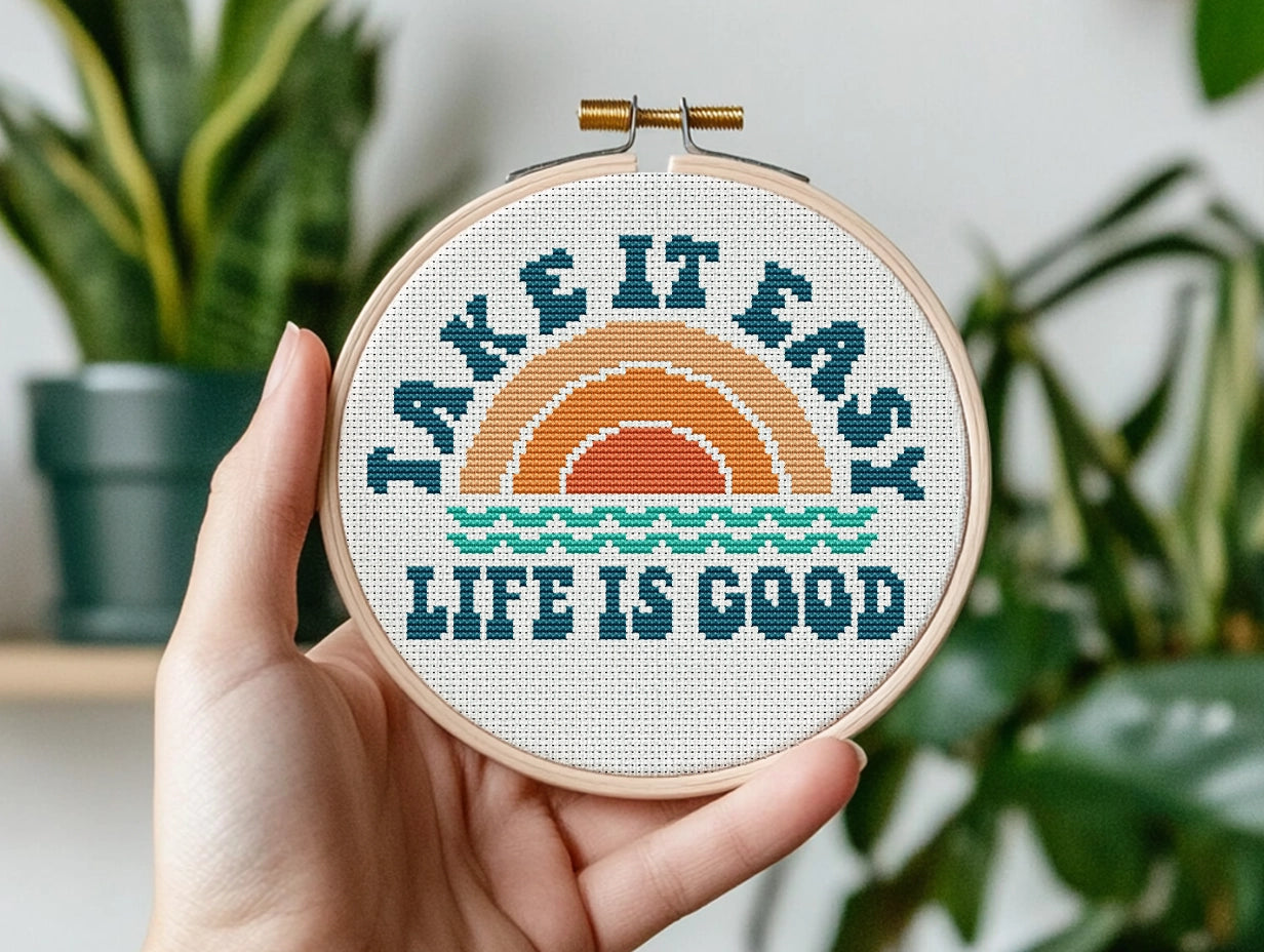 Embroidery hoop with 'Take it easy, life is good' text and sunset design held by a hand with plants in the background.