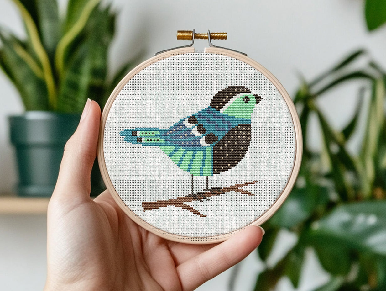 Hand holding a cross-stitched hoop with a colorful bird design against a plant background
