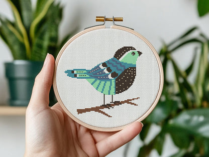 Hand holding a cross-stitched hoop with a colorful bird design against a plant background