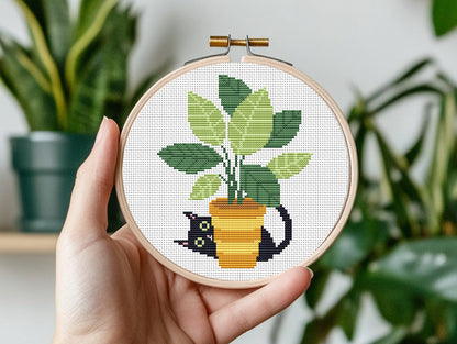Hand holding a cross-stitched hoop with a potted plant design, surrounded by green plants.