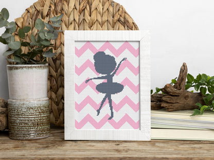 Framed cross-stitched art of a ballerina on pink chevron pattern with decorative items in the background.