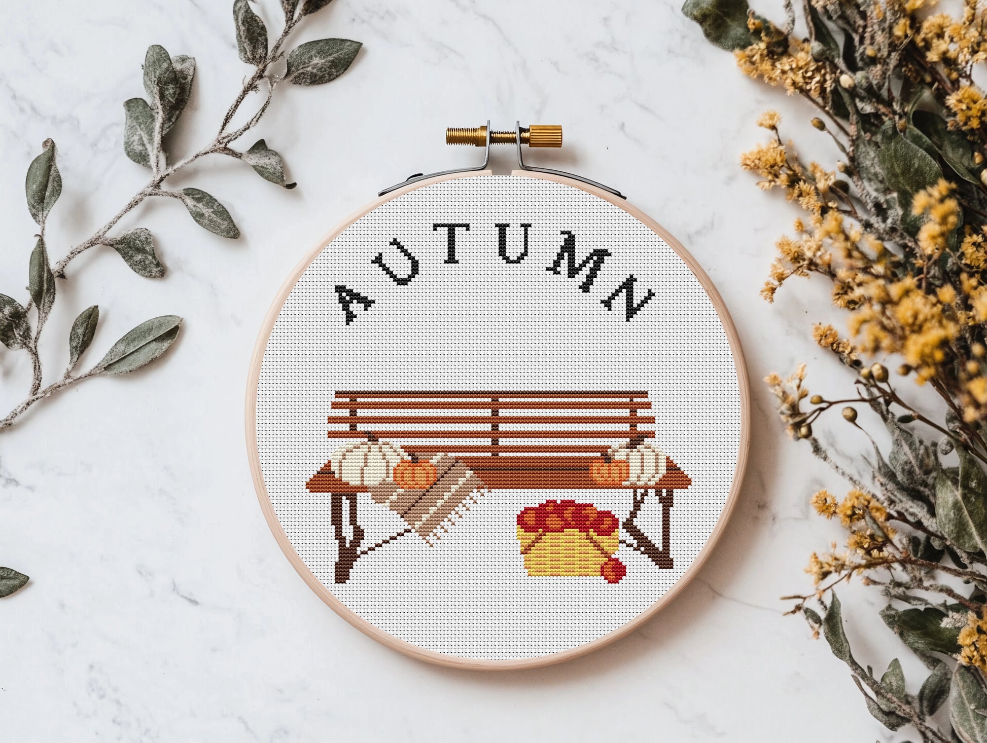 Cross-stitched hoop with 'AUTUMN' text and bench design on a white background with decorative elements.