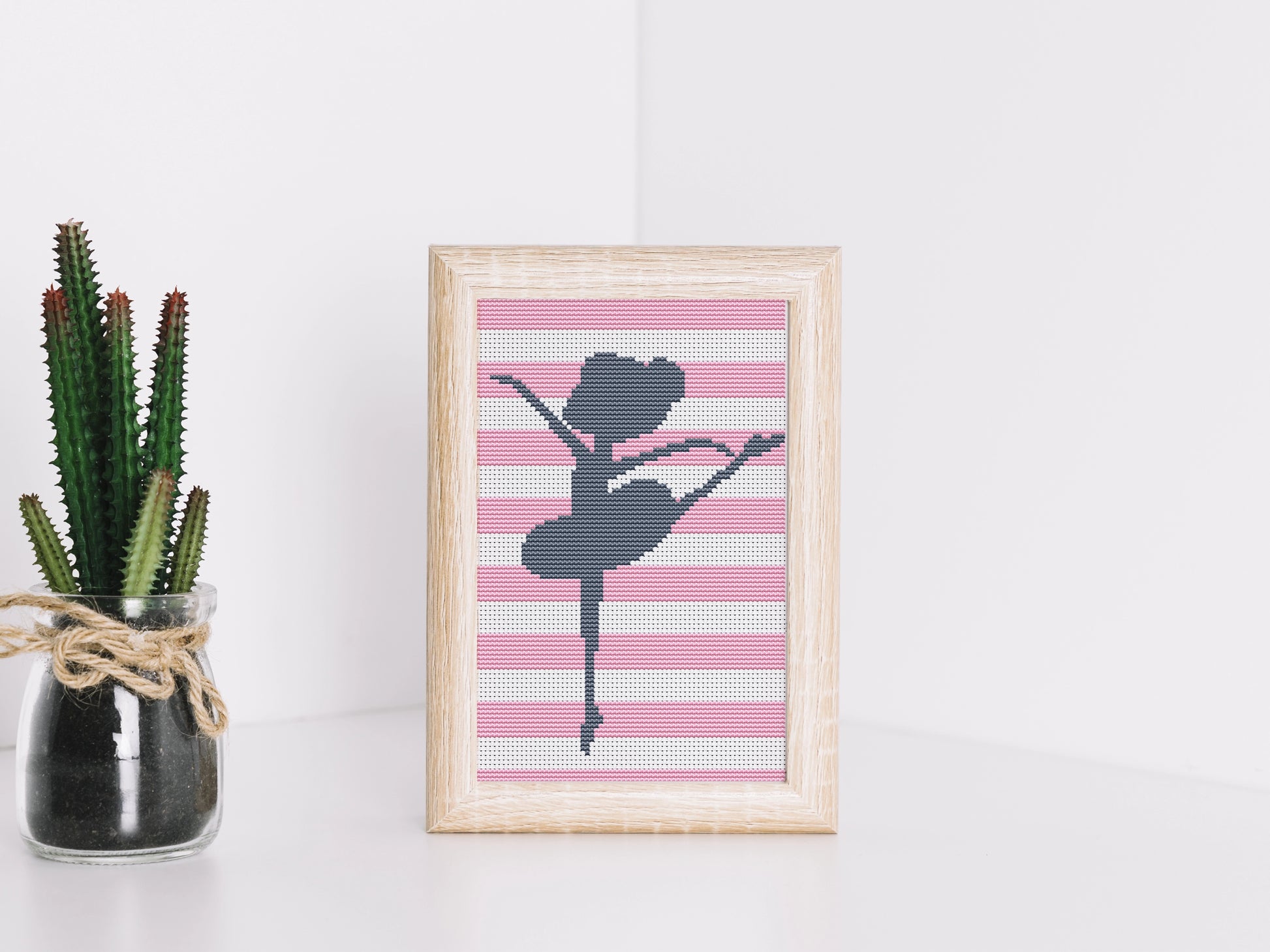 Framed cross-stitched art of a ballerina on a pink and white striped background, next to a potted plant.
