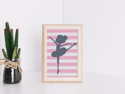 Framed cross-stitched art of a ballerina on a pink and white striped background, next to a potted plant.