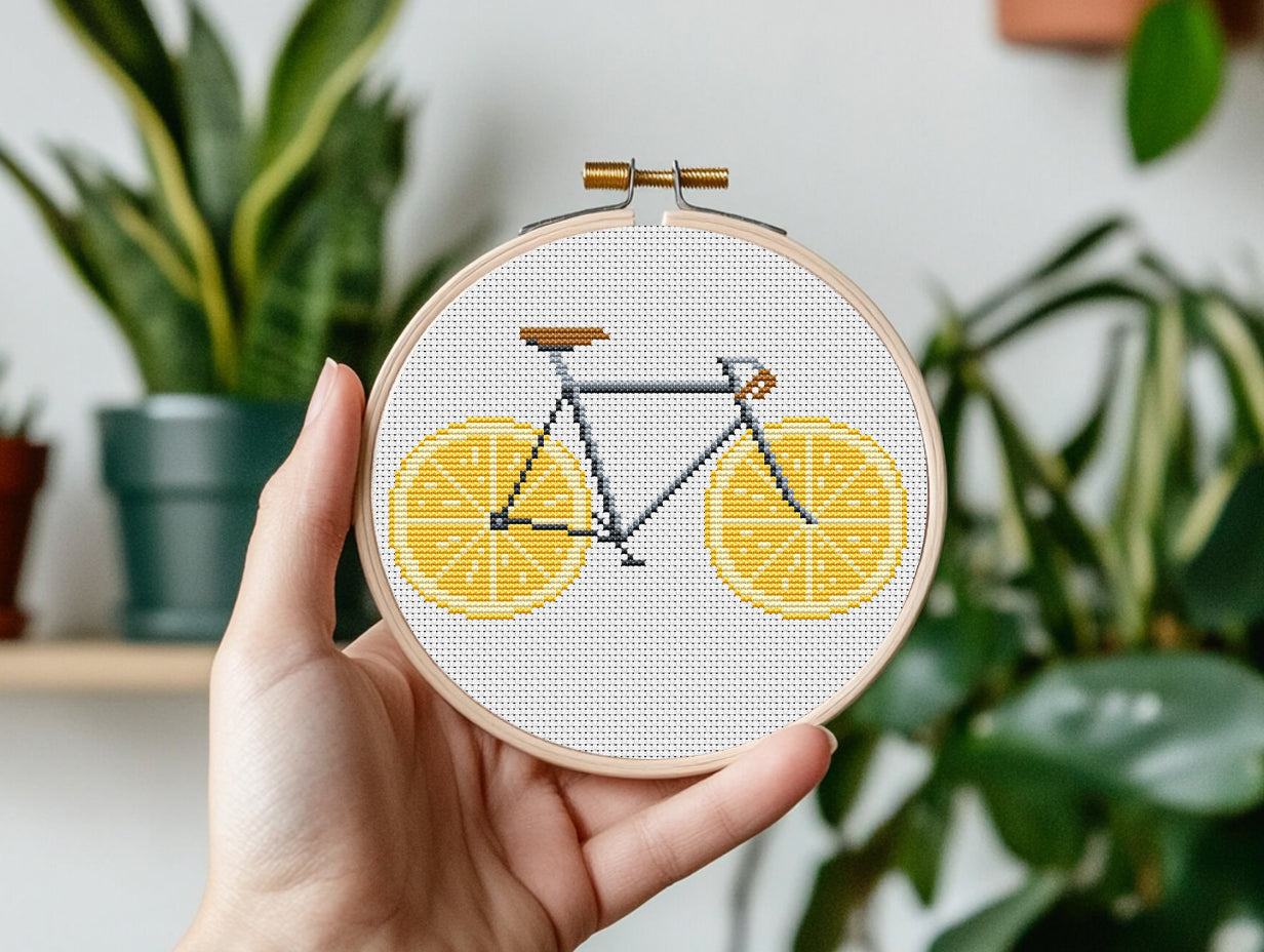 Hand holding a cross-stitched bicycle design with lemons in a hoop against a plant-filled background