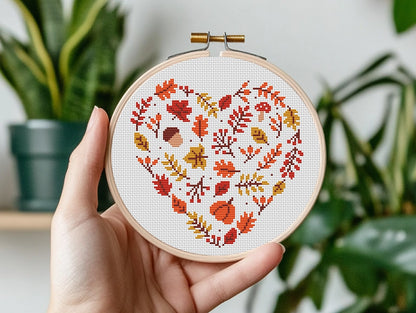 Hand holding a cross-stitched hoop with autumn leaf pattern against a blurred indoor plant background