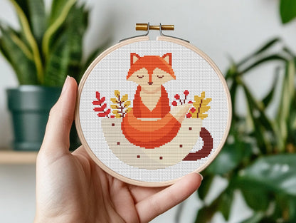 Hand holding a cross-stitched hoop with a fox design against a plant background