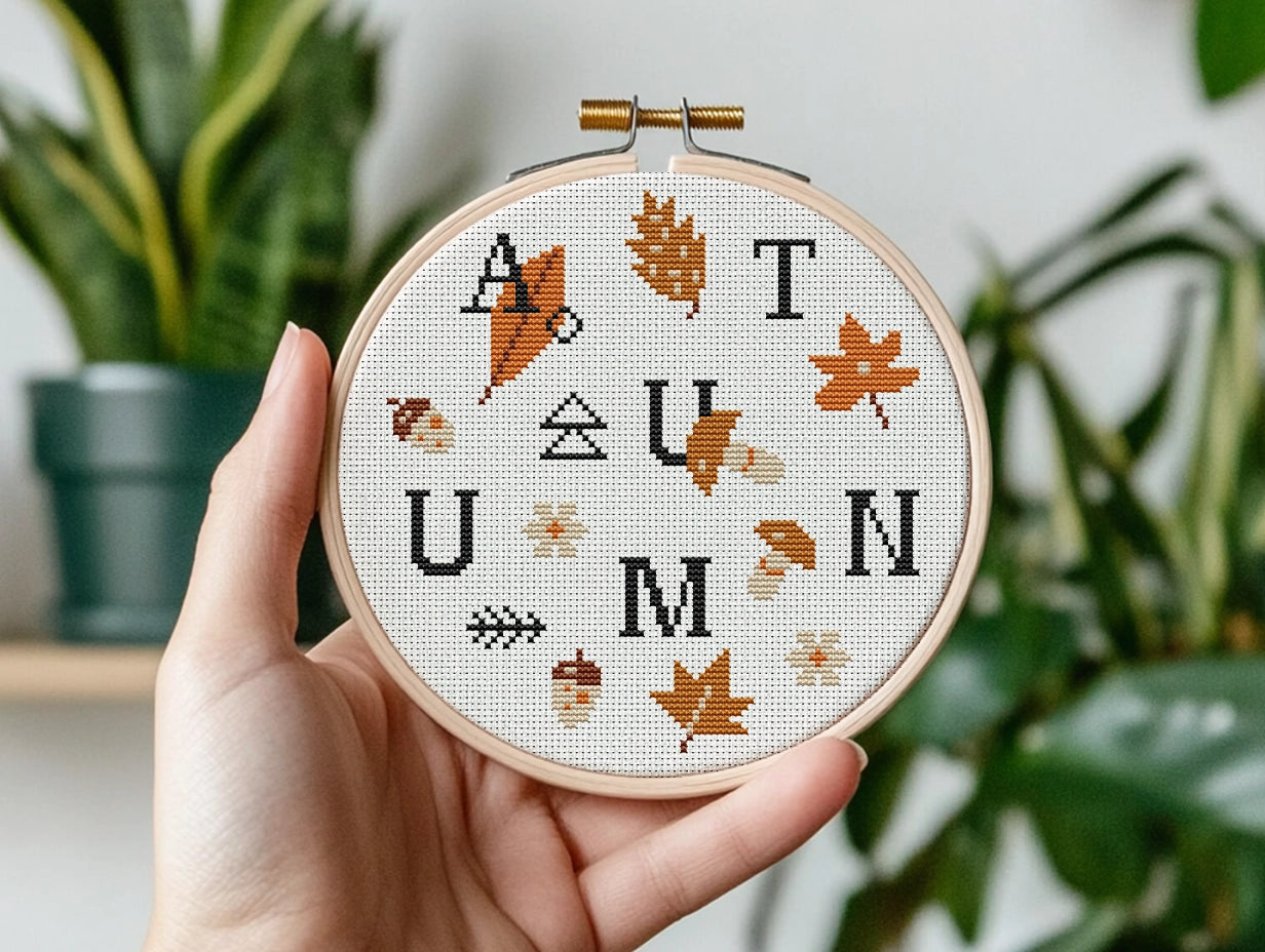 Embroidery hoop with 'AUTUMN' cross-stitch design held by a hand, with plants in the background.
