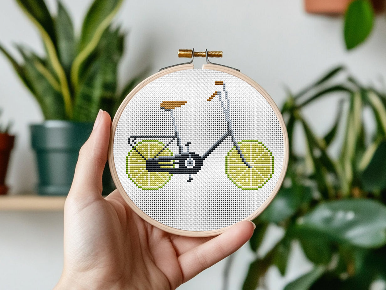 Hand holding a cross-stitched bicycle design with lime slices against a plant-filled background