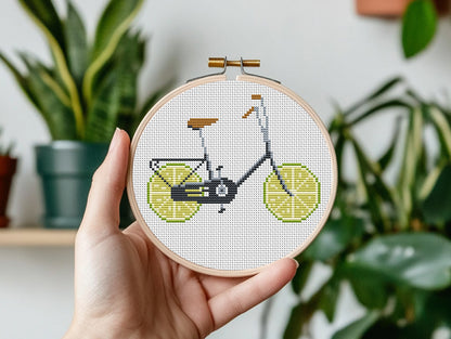 Hand holding a cross-stitched bicycle design with lime slices against a plant-filled background