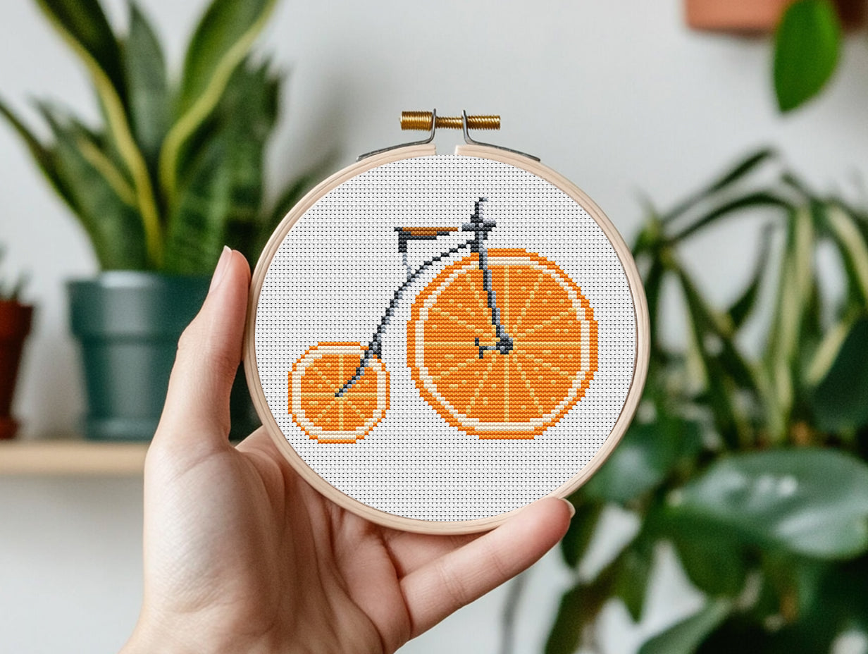 Hand holding a cross-stitched bicycle design on fabric with plants in the background