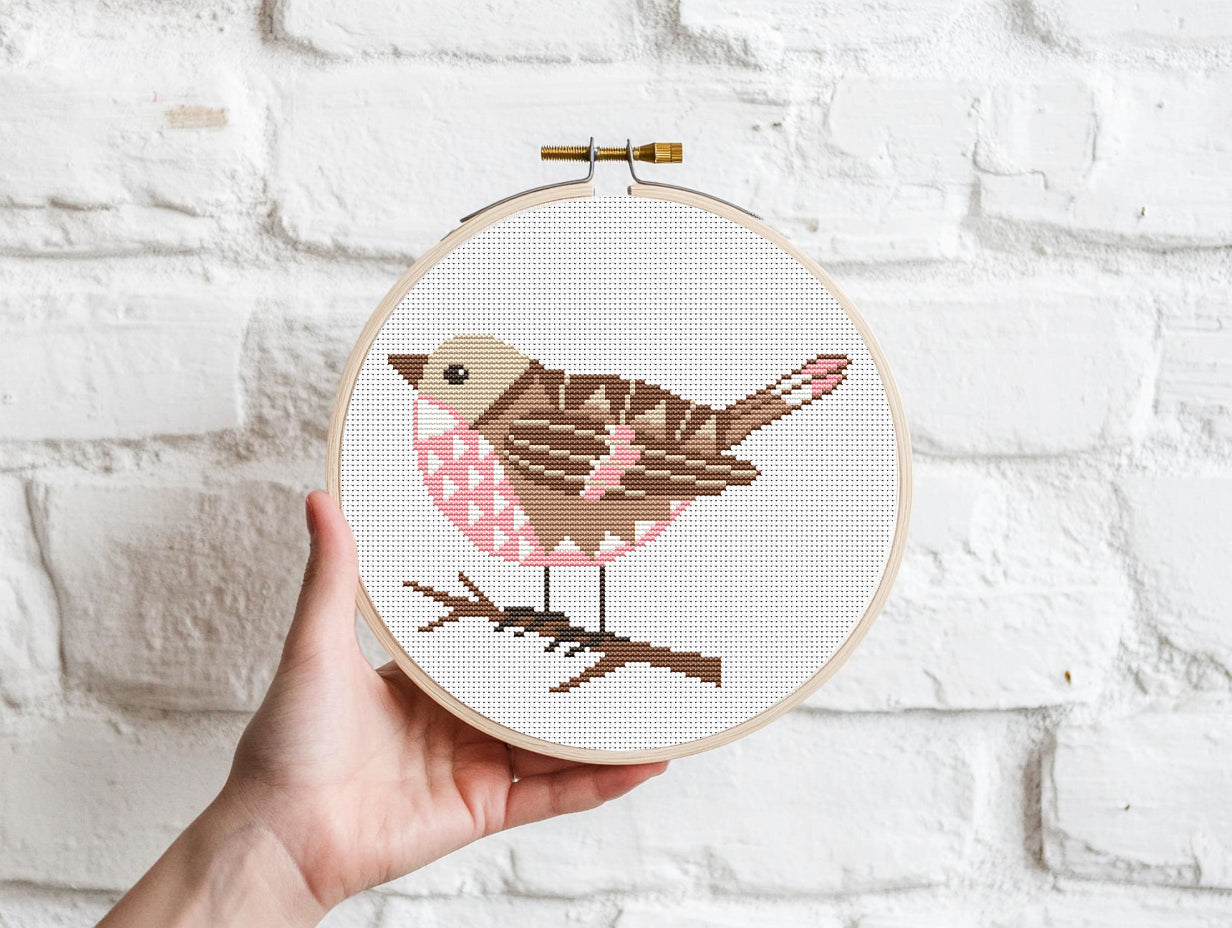 Hand holding a cross-stitched hoop with a bird design against a white brick wall.