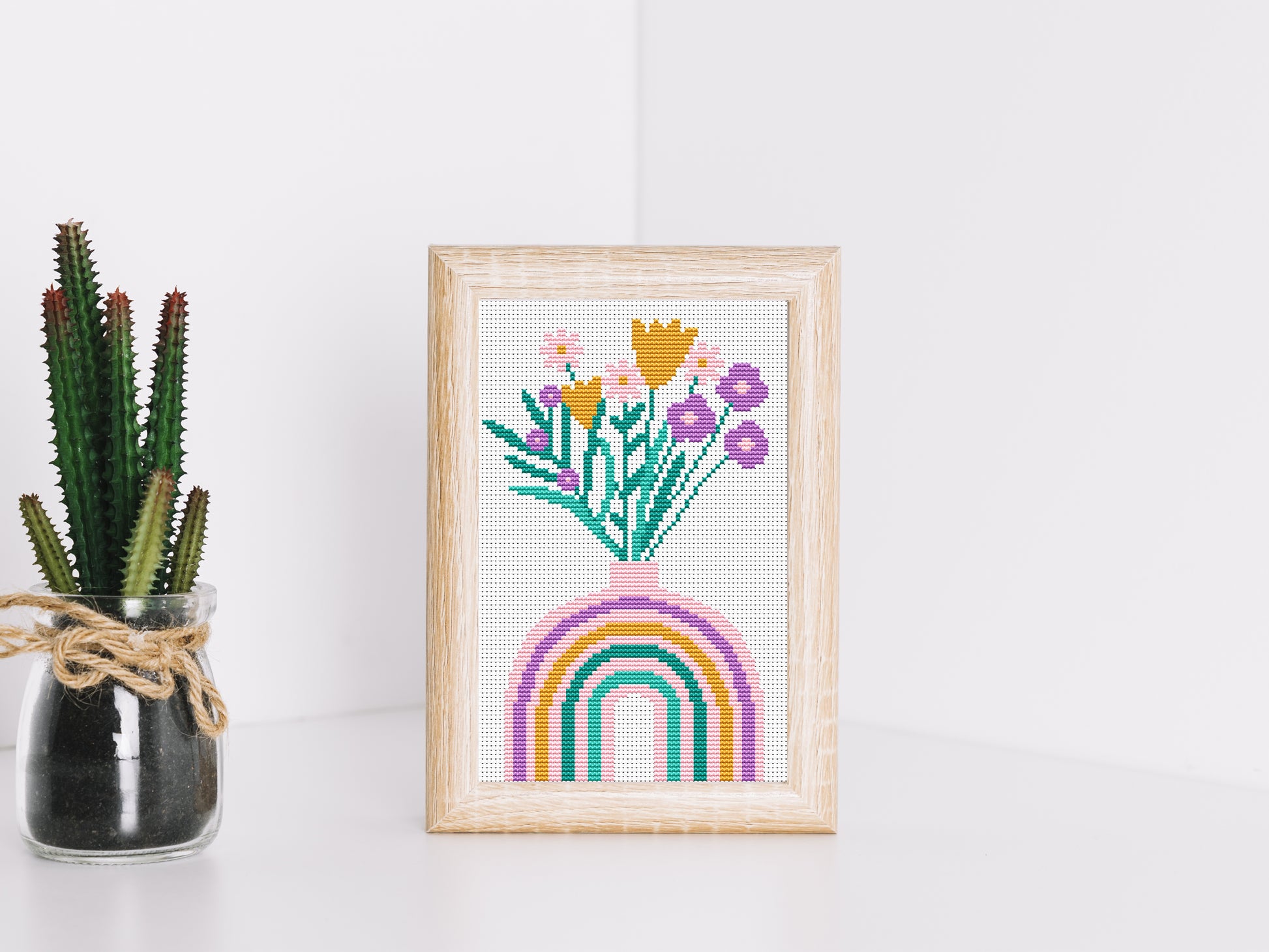 Framed cross-stitched artwork with flowers and a rainbow on a white surface with a plant.