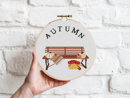 Cross-stitched hoop with 'AUTUMN' and bench design held against a white brick wall.