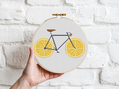 Embroidery hoop with a bicycle design made of yellow shapes held against a white brick wall.