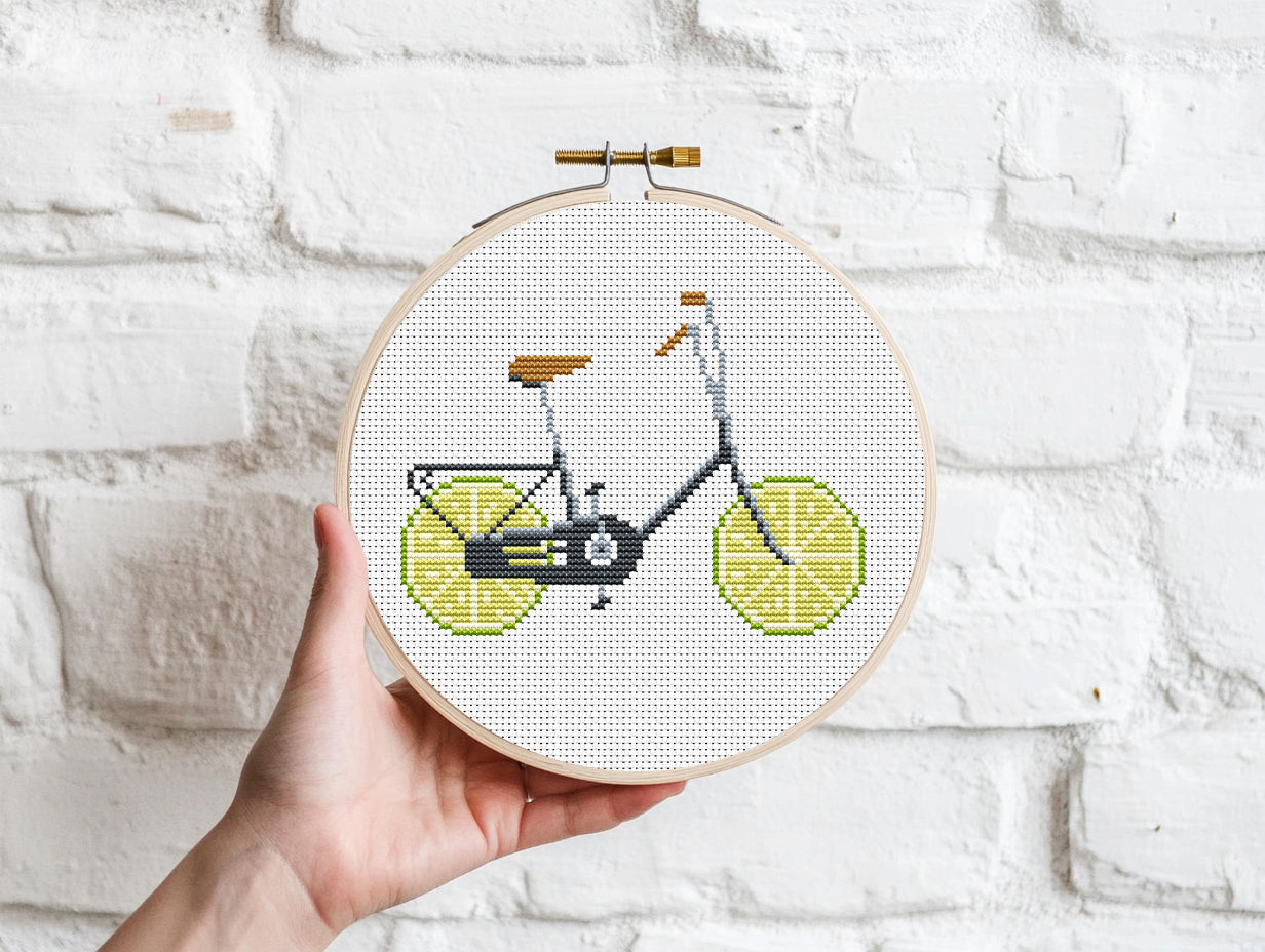 Embroidery hoop with a bicycle design made of lemons against a white brick wall.
