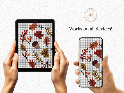 Hands holding a tablet and phone displaying cross-stitch patterns of leaves and acorns.